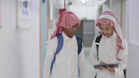 العمل الجماعي والتعاون بين الزملاء، مناقشة المواد الدراسيه، طالب عربي خليجي سعودي يرتدي الثوب الابيض و الشماغ و الحقيبة المدرسية يسير داخل المدرسة برفقة زميله ، استخدام الجهاز اللوحي فى الدراسة ، مفهوم التعليم الإلكتروني، استخدام الاجهزة التقنية الحديثة