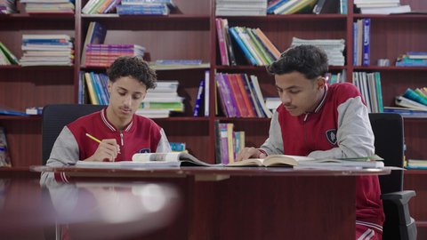حل التمارين الدراسية، طالبان عربيان سعوديان خليجييان يرتديان الزي المدرسي يجلسان في المكتبة المدرسية، الاستمتاع بقراءة الكتب المدرسية، التعليم في المملكة العربية السعودية