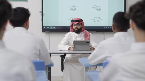 مفهوم الاجتهاد والتفوق، التعليم كوسيلة أساسية لتنمية القدرات العقلية وتوسيع الأفق الفكري, التعليم في المملكة العربية السعودية, اكتساب المعرفة و الخبرات العلمية, معلم عربي خليجي سعودي يرتدي الثوب التقليدي والشماغ يجلس على المكتب امامه التابلت يقوم بشرح الدروس للطلاب
