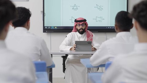 التعليم كوسيلة أساسية لتنمية القدرات العقلية وتوسيع الأفق الفكري, التعليم في المملكة العربية السعودية, اكتساب المعرفة و الخبرات العلمية, مفهوم الاجتهاد والتفوق, معلم عربي خليجي سعودي يرتدي الثوب التقليدي والشماغ يجلس على المكتب امامه التابلت يقوم بشرح الدروس للطلاب