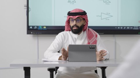 دور العلم في تحفيز العقل وتعزيز مهارات التحليل والفهم, التعليم في المملكة العربية السعودية, اكتساب المعرفة و الخبرات العلمية, مفهوم الاجتهاد والتفوق, معلم عربي خليجي سعودي يرتدي الثوب التقليدي والشماغ يجلس على المكتب امامه التابلت يقوم بشرح الدروس للطلاب