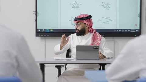 أهمية المعرفة في توسيع المدارك وتنمية التفكير العقلي, التعليم في المملكة العربية السعودية, اكتساب المعرفة و الخبرات العلمية, مفهوم الاجتهاد والتفوق, معلم عربي خليجي سعودي يرتدي الثوب التقليدي والشماغ يجلس على المكتب امامه التابلت يقوم بشرح الدروس للطلاب