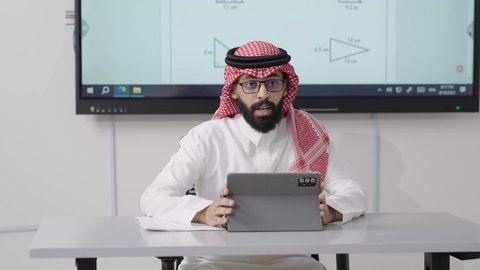 العلاقة بين التعلم وتطوير القدرات الذهنية والإبداعية, التعليم في المملكة العربية السعودية, اكتساب المعرفة و الخبرات العلمية, مفهوم الاجتهاد والتفوق, معلم عربي خليجي سعودي يرتدي الثوب التقليدي والشماغ يجلس على المكتب امامه التابلت يقوم بشرح الدروس للطلاب