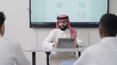 تعزيز القدرات الذهنية من خلال اكتساب المعرفة والتعلم, التعليم في المملكة العربية السعودية, اكتساب المعرفة و الخبرات العلمية, مفهوم الاجتهاد والتفوق, معلم عربي خليجي سعودي يرتدي الثوب التقليدي والشماغ يجلس على المكتب امامه التابلت يقوم بشرح الدروس للطلاب