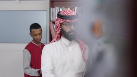 الاستمتاع بقراءة الكتب المدرسية، معلم عربي خليجي سعودي يرتدي الثوب الابيض و الشماغ يقف بجوار مكتبة خشبية في المكتبة المدرسية، ممارسة الهوايات المفضلة ، قضاء وقت ممتع ، التعليم في المملكة العربية السعودية