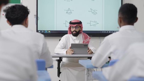 دور التعليم في تطوير التفكير وتنشيط القدرات الذهنية, التعليم في المملكة العربية السعودية, اكتساب المعرفة و الخبرات العلمية, مفهوم الاجتهاد والتفوق, معلم عربي خليجي سعودي يرتدي الثوب التقليدي والشماغ يجلس على المكتب امامه التابلت يقوم بشرح الدروس للطلاب
