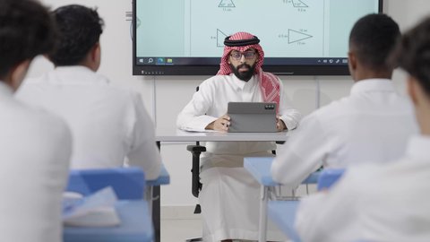 إدخال التقنيات الرقمية المتقدمة في أساليب التعليم الحديثة, التعليم في المملكة العربية السعودية, اكتساب المعرفة و الخبرات العلمية, مفهوم الاجتهاد والتفوق, معلم عربي خليجي سعودي يرتدي الثوب التقليدي والشماغ يجلس على المكتب امامه التابلت يقوم بشرح الدروس للطلاب