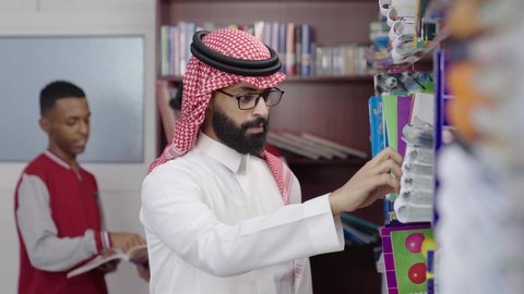 قضاء وقت ممتع ، ممارسة الهوايات المفضلة ، معلم عربي خليجي سعودي يرتدي الثوب الابيض و الشماغ يقف بجوار مكتبة خشبية في المكتبة المدرسية، الاستمتاع بقراءة الكتب المدرسية، التعليم في المملكة العربية السعودية