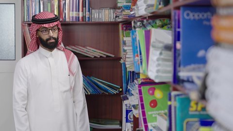 قضاء وقت ممتع ، ممارسة الهوايات المفضلة ، معلم عربي خليجي سعودي يرتدي الثوب الابيض و الشماغ يقف بجوار مكتبة خشبية في المكتبة المدرسية، الاستمتاع بقراءة الكتب المدرسية، التعليم في المملكة العربية السعودية