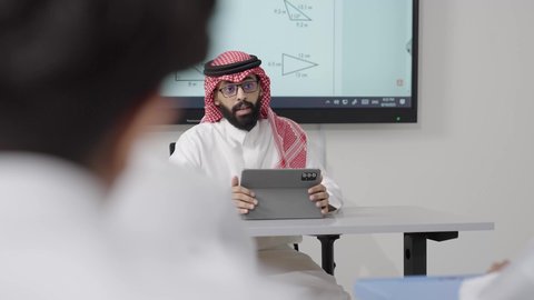 استخدام التقنيات الرقمية لتعزيز العملية التعليمية, التعليم في المملكة العربية السعودية, اكتساب المعرفة و الخبرات العلمية, مفهوم الاجتهاد والتفوق, معلم عربي خليجي سعودي يرتدي الثوب التقليدي والشماغ يجلس على المكتب امامه التابلت يقوم بشرح الدروس للطلاب