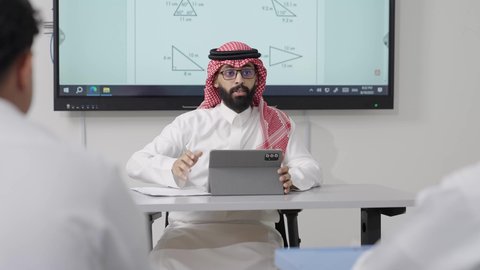 دمج التقنيات الحديثة والمتطورة مع العملية التعليمية, التعليم في المملكة العربية السعودية, اكتساب المعرفة و الخبرات العلمية, مفهوم الاجتهاد والتفوق, معلم عربي خليجي سعودي يرتدي الثوب التقليدي والشماغ يجلس على المكتب امامه التابلت يقوم بشرح الدروس للطلاب