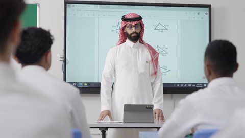 Supporting students' skills through innovative teaching methods, using modern technologies, an Arab Gulf Saudi math teacher wearing traditional attire and a headscarf places a tablet in front of him and explains mathematical problems on an electronic screen, the modern and developed curricula and educational programs in the Kingdom of Saudi Arabia.