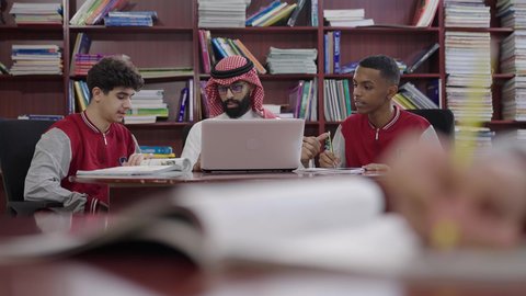 جودة التعليم في السعودية، المشاركة الفعالة للطلاب مع المدرسين، حوارات ونقاشات بين الاستاذة والطلاب، معلم عربي خليجي سعودي يرتدي الثوب التقليدي والشماغ يجلس امام الحاسب المحمول يقوم بمناقشة الدروس مع طلابه داخل المكتبة المدرسية، التربية والتعليم بالمناهج المقررة للتلاميذ، العودة إلى المدارس