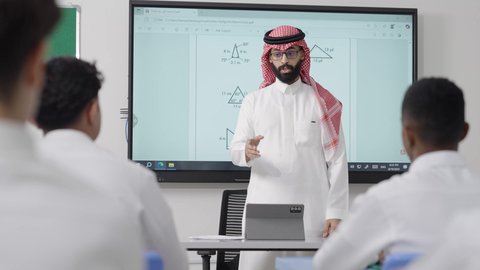 Enhancing students' capabilities through interactive education, using modern technologies, an Arab Gulf Saudi math teacher wearing traditional attire and a headscarf places a tablet in front of him and explains mathematical problems on an electronic screen, the modern and developed curricula and educational programs in the Kingdom of Saudi Arabia.