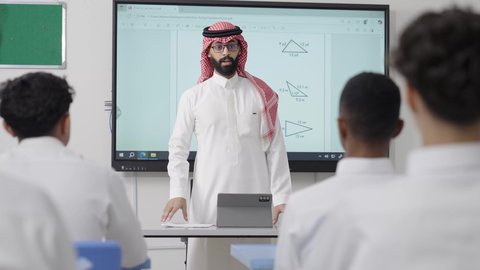 Raising hands to answer the question, an Arab Gulf Saudi math teacher wearing traditional attire and a headscarf has a tablet in front of him and is explaining mathematical problems on an electronic screen, the modern and developed curricula and educational programs in the Kingdom of Saudi Arabia, focusing on students' abilities by delivering lessons, using modern technologies.