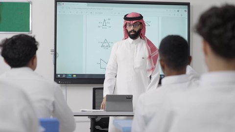 A Gulf Arab Saudi math teacher wearing traditional attire and a shemagh has a tablet in front of him and is explaining mathematical problems on an electronic screen, focusing on modern and developed educational curricula in the Kingdom of Saudi Arabia, paying attention to students' abilities by delivering lessons, and using modern technologies.