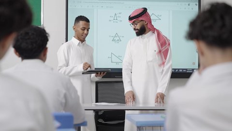 التفاعل والنشاط داخل الفصل الدراسي، استخدام التقنية الحديثة، طالب عربي خليجي سعودي يرتدي الثوب الابيض يمسك بيديه جهاز التابلت و يقوم بشرح الدرس برفقة معلمه داخل الفصل، أهداف التربية والتعليم بالمناهج المقررة للطلاب والطالبات في السعودية