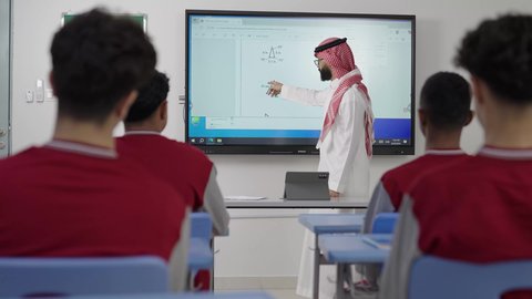 المقررات والمناهج التعليمية الحديثة والمطورة بالمملكة العربية السعودية، الاهتمام بقدرات الطلاب بتقديم الدروس, معلم رياضيات عربي خليجي سعودي يرتدي الثوب التقليدي والشماغ يضع امامه التابلت يقوم بشرح المسائل الحسابية على  شاشة الكترونية، استخدام التقنيات الحديثة