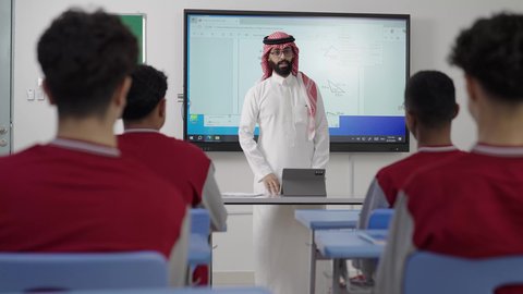 Focusing on students' abilities by delivering lessons, an Arab Gulf Saudi math teacher wearing traditional attire and a headscarf places a tablet in front of him and explains mathematical problems on an electronic screen, the modern and developed curricula and educational programs in the Kingdom of Saudi Arabia, using modern technologies.