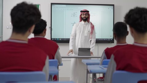 Two Saudi Gulf Arab students are sitting in their seats, and in front of them is a Saudi teacher using a tablet for explanation. Active participation in class discussions, benefiting from advanced technologies in learning and studying, the evolution of teaching methods in schools in the Kingdom, developed curricula, and e-learning.