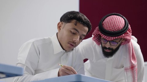 دور التعليم في تطوير التفكير والقدرات الذهنية، مفهوم الاجتهاد وطلب التفوق، التربية والتعليم في السعودية، معلم عربي خليجي سعودي يرتدي الثوب الابيض و الشماغ يقوم بمساعدة طالب اثناء تقديم الامتحانات