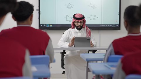 Active participation in classroom discussions, benefiting from advanced technologies in learning and studying, the evolution of teaching methods in the schools of the Kingdom, developed curricula and e-learning, Gulf Arab Saudi students sitting in their seats with a Saudi teacher using a tablet for explanation.