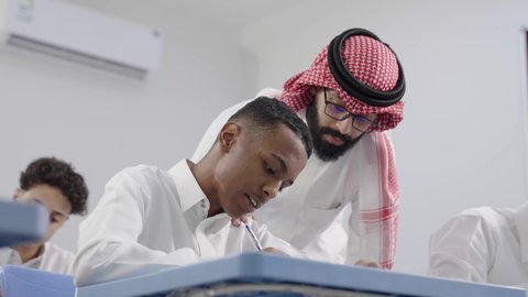 مفهوم الاجتهاد وطلب التفوق، أهمية العلم في تنمية القدرات العقلية، التربية والتعليم في السعودية، معلم عربي خليجي سعودي يرتدي الثوب الابيض و الشماغ يقوم بمساعدة طالب اثناء تقديم الامتحانات