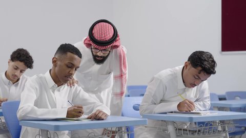 ايماءات التمعن والتركيز، أهمية العلم في تنمية القدرات العقلية، التربية والتعليم في السعودية، معلم عربي خليجي سعودي يرتدي الثوب الابيض و الشماغ  يقوم بمساعدة الطلاب اثناء تقديم الامتحانات، مفهوم الاجتهاد وطلب التفوق