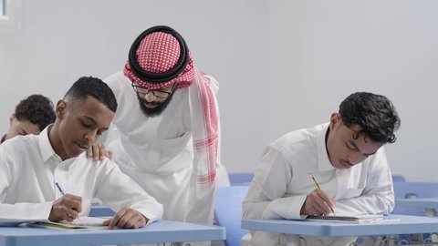 أهمية العلم في تنمية القدرات العقلية، ايماءات التمعن والتركيز، التربية والتعليم في السعودية، معلم عربي خليجي سعودي يرتدي الثوب الابيض و الشماغ  يقوم بمساعدة الطلاب اثناء تقديم الامتحانات، مفهوم الاجتهاد وطلب التفوق