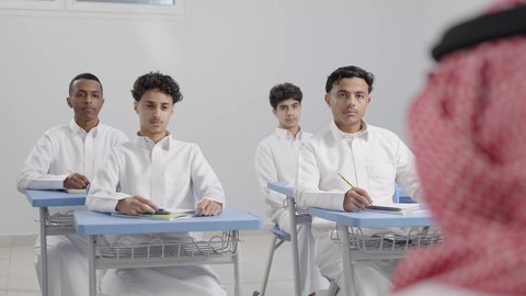 المشاركة الفعالة للطلاب مع المدرسين، تصوير مقرب من الخلف لمعلم عربي خليجي سعودي يرتدي الشماغ يقوم بمناقشة الدروس مع طلابه، جودة التعليم في السعودية، التربية والتعليم بالمناهج المقررة للتلاميذ، حوارات ودية بين الاستاذ والطلاب