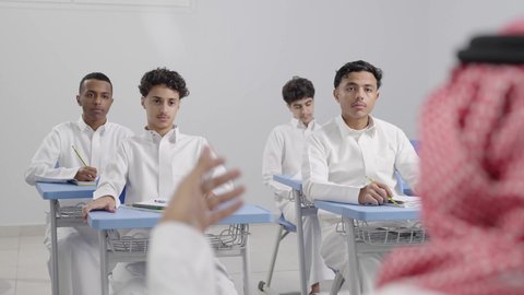 المشاركة الفعالة للطلاب مع المدرسين، تصوير مقرب من الخلف لمعلم عربي خليجي سعودي يرتدي الشماغ يقوم بمناقشة الدروس مع طلابه، جودة التعليم في السعودية، التربية والتعليم بالمناهج المقررة للتلاميذ، حوارات ودية بين الاستاذ والطلاب