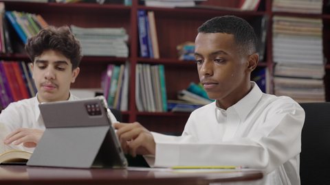 مراجعة الدروس اليوميه، حل الواجبات المدرسية عبر التابلت، التعليم في المملكة العربية السعودية، استخدام التقنية الحديثة، الاستمتاع بقراءة الكتب المدرسية، تصوير مقرب لطالبان عربيان خليجيان سعوديان يرتديان الثوب الابيض يجلسان في المكتبة المدرسية