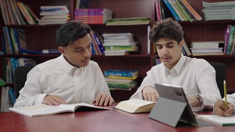 Group study and collaboration among colleagues, completing homework assignments, using modern technology, enjoying reading school books, a close-up shot of two Arab Gulf Saudi students wearing white thobes sitting in the school library, education in the Kingdom of Saudi Arabia.