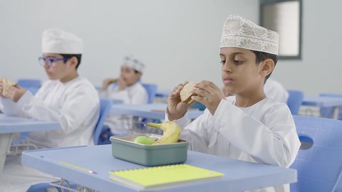 موسم العودة للمدارس، تناول الأطعمة الصحية في وقت الفسحة المدرسية، الوجبات والسناكات الغنية بالعناصر الغذائية الهامة لبناء الجسم، مجموعة من الطلاب العربيين الخليجيين العمانيين يرتدون الدشداشة والكمة يجلسون في مقاعدهم وأمامهم اللانش البوكس