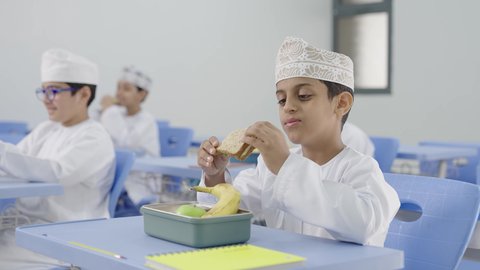 تناول الأطعمة الصحية في وقت الفسحة المدرسية، موسم العودة للمدارس، الوجبات والسناكات الغنية بالعناصر الغذائية الهامة لبناء الجسم، مجموعة من الطلاب العربيين الخليجيين العمانيين يرتدون الدشداشة والكمة يجلسون في مقاعدهم وأمامهم اللانش البوكس