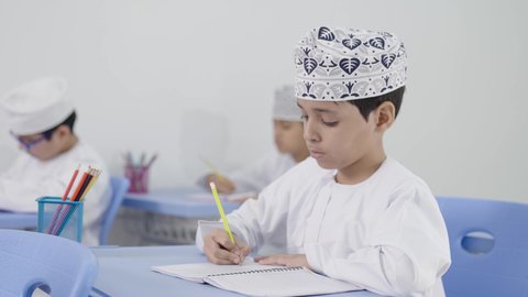 العودة إلى المدارس،تأدية الواجبات المدرسية، الإستعداد للعام الدراسي الجديد، التعليم في سلطنة عمان،  مجموعة من الطلاب العربيين الخليجيين العمانيين يرتدون الدشداشة والكمة يجلسون في مقاعدهم ويقومون بحل الواجب
