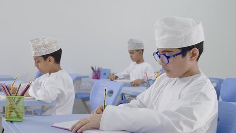 تأدية الواجبات المدرسية، الإستعداد للعام الدراسي الجديد، التعليم في سلطنة عمان، العودة إلى المدارس، مجموعة من الطلاب العربيين الخليجيين العمانيين يرتدون الدشداشة والكمة يجلسون في مقاعدهم ويقومون بحل الواجب