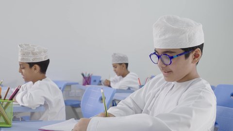 A group of Arab Gulf Omani students wearing dishdasha and kumma are sitting in their seats and doing their homework, completing school assignments, preparing for the new school year, education in the Sultanate of Oman, back to school.