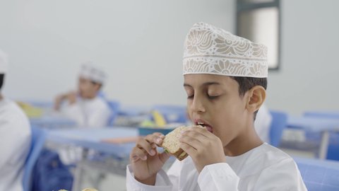 تناول الأطعمة الصحية في وقت الفسحة المدرسية، موسم العودة للمدارس، الوجبات والسناكات الغنية بالعناصر الغذائية الهامة لبناء الجسم، مجموعة من الطلاب العربيين الخليجيين العمانيين يرتدون الدشداشة والكمة يجلسون في مقاعدهم وأمامهم اللانش البوكس
