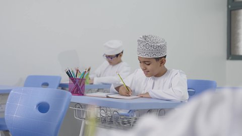 الإستعداد للعام الدراسي الجديد، تأدية الواجبات المدرسية، التعليم في سلطنة عمان، العودة إلى المدارس، مجموعة من الطلاب العربيين الخليجيين العمانيين يرتدون الدشداشة والكمة يجلسون في مقاعدهم ويقومون بحل الواجب