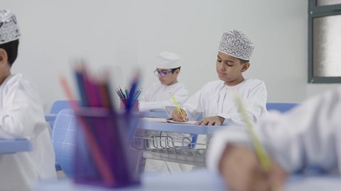 تأدية الواجبات المدرسية، الإستعداد للعام الدراسي الجديد، التعليم في سلطنة عمان، العودة إلى المدارس، مجموعة من الطلاب العربيين الخليجيين العمانيين يرتدون الدشداشة والكمة يجلسون في مقاعدهم ويقومون بحل الواجب
