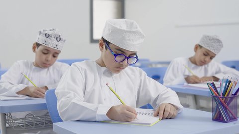 مجموعة من الطلاب العربيين الخليجيين العمانيين يرتدون الدشداشة والكمة يجلسون في مقاعدهم ويقومون بحل الواجب ، تأدية الواجبات المدرسية، الإستعداد للعام الدراسي الجديد، التعليم في سلطنة عمان، العودة إلى المدارس