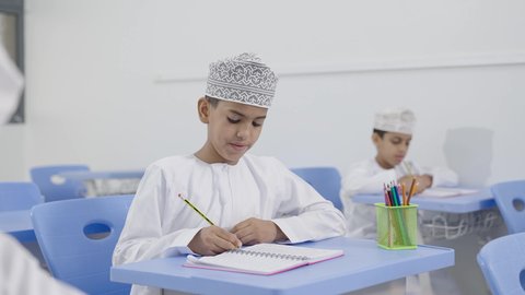 العودة إلى المدارس،تأدية الواجبات المدرسية، الإستعداد للعام الدراسي الجديد، التعليم في سلطنة عمان،  مجموعة من الطلاب العربيين الخليجيين العمانيين يرتدون الدشداشة والكمة يجلسون في مقاعدهم ويقومون بحل الواجب