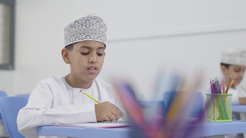 التعليم في سلطنة عمان،تأدية الواجبات المدرسية، الإستعداد للعام الدراسي الجديد، العودة إلى المدارس، مجموعة من الطلاب العربيين الخليجيين العمانيين يرتدون الدشداشة والكمة يجلسون في مقاعدهم ويقومون بحل الواجب