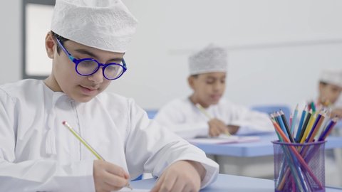 العودة إلى المدارس،تأدية الواجبات المدرسية، الإستعداد للعام الدراسي الجديد، التعليم في سلطنة عمان،  مجموعة من الطلاب العربيين الخليجيين العمانيين يرتدون الدشداشة والكمة يجلسون في مقاعدهم ويقومون بحل الواجب