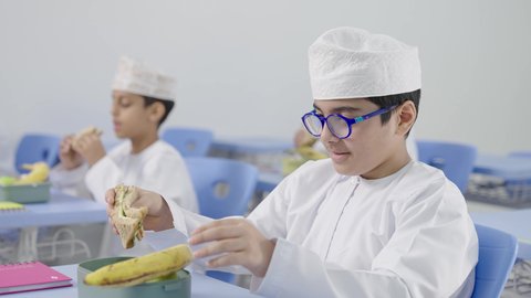 تناول الأطعمة الصحية في وقت الفسحة المدرسية، موسم العودة للمدارس، الوجبات والسناكات الغنية بالعناصر الغذائية الهامة لبناء الجسم، مجموعة من الطلاب العربيين الخليجيين العمانيين يرتدون الدشداشة والكمة يجلسون في مقاعدهم وأمامهم اللانش البوكس