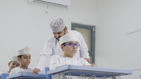 معلم عربي خليجي عماني يرتدي الدشداشة والكمة يقوم بمناقشة الدروس مع طلابه، جودة التعليم في سلطنة عمان ، التربية والتعليم بالمناهج المقررة للتلاميذ،المشاركة الفعالة للطلاب مع المدرسين، حوارات ودية بين الاستاذ والطلاب