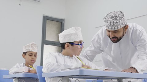 Active participation of students with teachers, a Gulf Arab Omani teacher wearing a dishdasha and kumma discussing lessons with his students, the quality of education in the Sultanate of Oman, education and teaching with the prescribed curricula for students, friendly dialogues between the teacher and students.