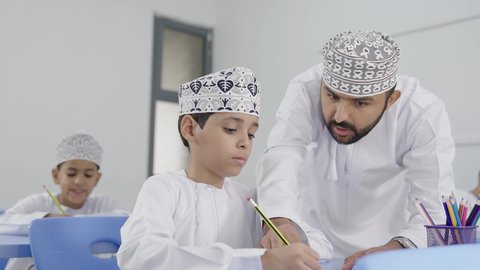 معلم عربي خليجي عماني يرتدي الدشداشة والكمة يقوم بمناقشة الدروس مع طلابه،المشاركة الفعالة للطلاب مع المدرسين،  جودة التعليم في سلطنة عمان ، التربية والتعليم بالمناهج المقررة للتلاميذ، حوارات ودية بين الاستاذ والطلاب