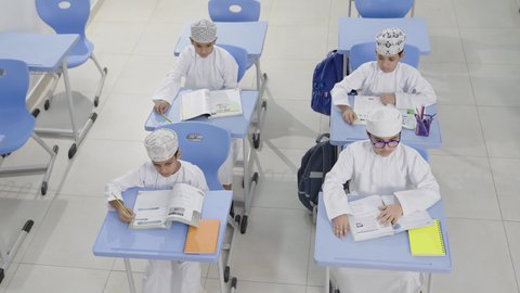 مفهوم التعليم والثقافة ، تأدية الواجبات المدرسية، الإستعداد للعام الدراسي الجديد، التعليم في سلطنة عمان، العودة إلى المدارس، مجموعة من الطلاب العربيين الخليجيين العمانيين يرتدون الدشداشة والكمة يجلسون في مقاعدهم ويقومون بحل الواجب